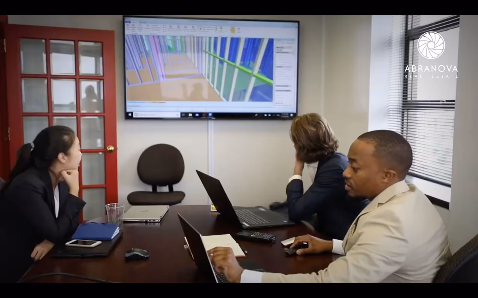 Three colleagues at a boardroom table reviewing a site map on a wall-mounted screen bearing the Abranova mark.